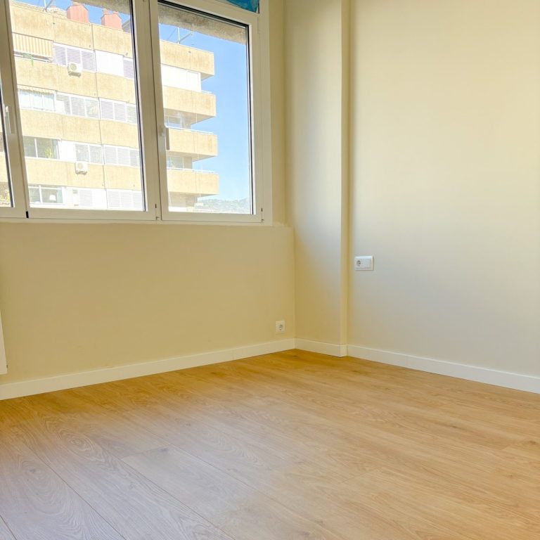 Habitación vacía con paredes pintadas beige y suelo de madera, ventana con acceso a balcón.