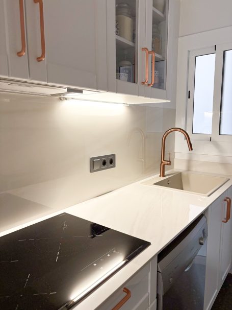 Cocina moderna con encimera blanca, fregadero, horno y detalles en metal cobre.