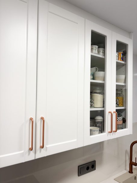 Muebles de cocina blancos con puertas de vidrio y tiradores de cobre.