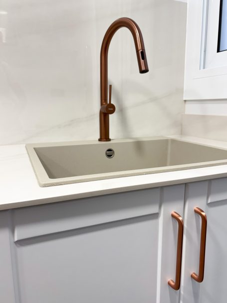 Grifo de cocina moderno de color cobre sobre un fregadero beige con muebles blancos.